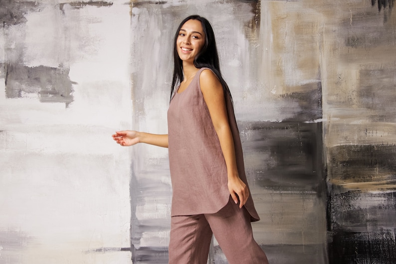Op de afbeelding: Een vrouw draagt een loszittende, mouwloze linnen jumpsuit in een stoffige roze kleur. De jumpsuit heeft een rechte pijp en een licht afgeronde zoom. De vrouw staat voor een getextureerde muur met een neutrale kleurenpalet.