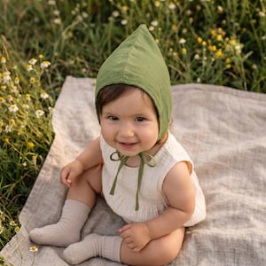 Può includere: Un bambino che indossa un cappuccio verde con la parte superiore a punta e un body senza maniche color crema. Il cappuccio ha un fiocco verde abbinato. Il bambino indossa anche calzini beige ed è seduto su una coperta di colore neutro.
