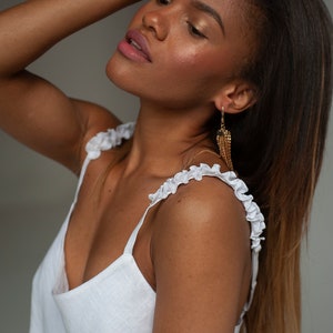 Pu&ograve; includere: Una donna con lunghi capelli castani indossa un top bianco in lino con spalline arricciate e orecchini pendenti dorati.