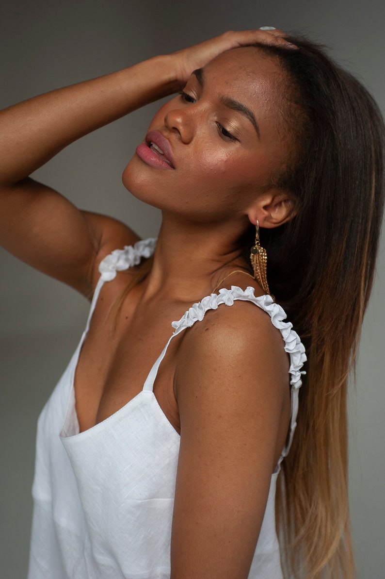 Pu&ograve; includere: Una donna con lunghi capelli castani indossa un top bianco in lino con spalline arricciate e orecchini pendenti dorati.