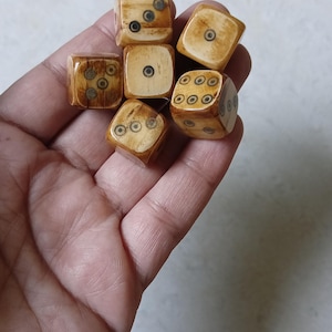 May include: A collection of seven antique dice, each with a light brown, aged appearance. The dice feature dark circular markings indicating the numbers, with varying arrangements of dots on each face. The dice are held in a hand.