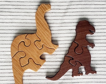 May include: Two wooden dinosaur puzzles. One is a light brown Brontosaurus, and the other is a dark brown T-Rex. Each puzzle is made of several interlocking pieces. The puzzles are on a white and gray striped surface.