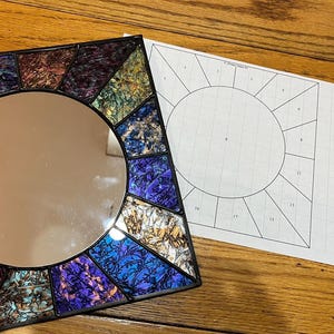 May include: A square mirror with a round center, surrounded by colorful stained glass in shades of blue, purple, and gold. A paper with a sunburst design is next to the mirror. The mirror is on a wooden surface.
