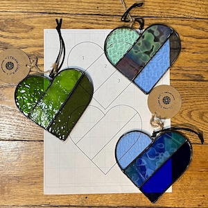 Puede incluir: Tres adornos de corazón de vidrieras en tonos verdes y azules, cada uno con un borde negro y una cuerda para colgar. Se ve un patrón de papel con contornos de corazón. Los adornos están sobre una superficie de madera.