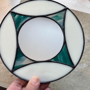 May include: A round stained glass piece with a white center and outer ring. The design includes four green and white sections, framed by black lines. The glass has a textured appearance, and the piece is held by a hand.