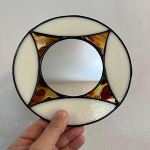 May include: Round stained glass mirror with a black frame. The mirror features cream-colored and amber-colored glass pieces. The mirror is held by a hand.