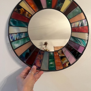 May include: Round stained glass mirror with a black metal frame. The colorful glass pieces are arranged in a radial pattern, featuring shades of red, orange, yellow, green, and purple. The mirror is held by a hand.