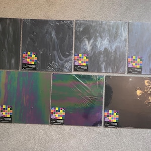 May include: Seven square sheets of stained glass arranged in two rows. The sheets feature various textures and colors, including black, gray, blue, and iridescent finishes. Each sheet has a small, colorful square sticker in the bottom corner.