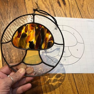 May include: A stained glass mushroom with a black metal frame and a hanging loop. The mushroom cap features shades of orange, yellow, and brown. The stem is yellow. A pattern for the mushroom is visible in the background.