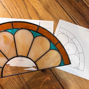 Puede incluir: Un vitral parcialmente terminado con un diseño en forma de abanico. El vidrio presenta tonos de naranja, amarillo y turquesa, enmarcado por líneas negras. Una guía de patrones numerada se encuentra junto a la obra, sugiriendo un proyecto de manualidades.