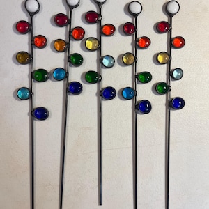 May include: Five decorative metal garden stakes with colorful glass orbs. Each stake has a white orb at the top, followed by a series of red, orange, yellow, green, and blue orbs. The stakes are arranged vertically against a light background.