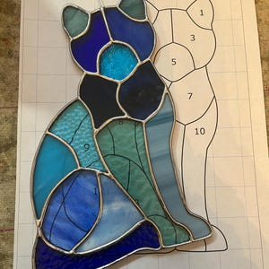 Op de afbeelding: Een gebrandschilderde kattenfiguur in blauwe, teal en turkooizen tinten, met een zilveren metalen omtrek. De kat zit, en het ontwerp staat op een geruit papier met een bijpassende omtrek. De afbeelding bevat de tekst "Jthrees Glass Art."