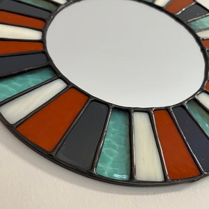 May include: A round mirror with a stained glass frame. The frame is made of black, white, orange, and teal glass pieces.