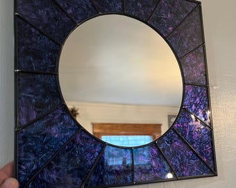 May include: Square mirror with a circular center. The frame is black, and the surrounding panels are a mix of purple and blue stained glass. The mirror is mounted on a white wall.