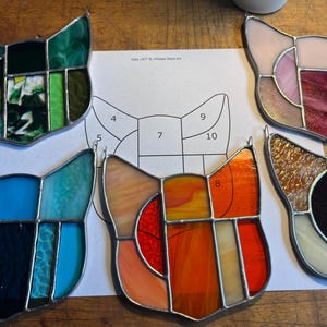 May include: Four cat-shaped stained glass suncatchers in green, blue, orange, and pink tones, arranged around a central paper pattern labeled Kitty Cat J.P. Glass Art with numbered sections for assembly and construction.