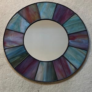 May include: Round decorative mirror with a stained glass design. The mirror's frame is composed of segments of glass in shades of purple, blue, and green. The mirror has a black metal frame and a reflective center.