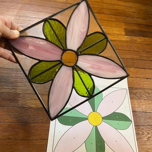May include: Stained glass flower art piece with pink petals, green leaves, and a yellow centre, set in a black metal frame. The artwork is held up, with a matching pattern on a paper below.