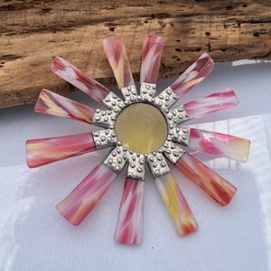 May include: A pink and yellow glass flower brooch with a silver center. The flower has a textured center and is surrounded by 12 petals.