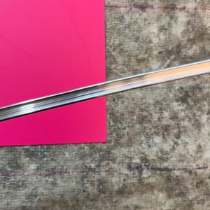 May include: A shiny, silver metal strip, possibly a decorative trim or molding, is positioned diagonally across a vibrant pink surface. The background features a textured, gray surface with subtle markings.