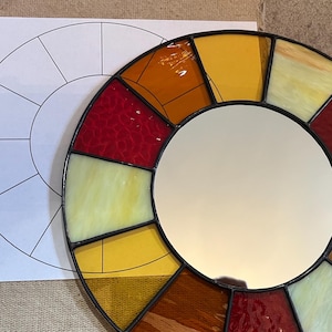 May include: A round stained glass mirror with a central reflective surface. The mirror is surrounded by a mosaic of colorful glass pieces in shades of red, yellow, and orange. A paper with a circular design is in the background.