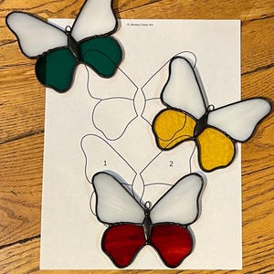 Puede incluir: Tres adornos de mariposa de vidrieras con alas blancas y cuerpos coloridos en verde, amarillo y rojo. Las mariposas están sobre un papel blanco con un contorno de mariposa. El fondo es una superficie de madera.