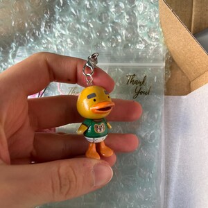 May include: A yellow duck character keychain with a green shirt featuring a brown bear graphic. The duck has a black beak and orange feet.