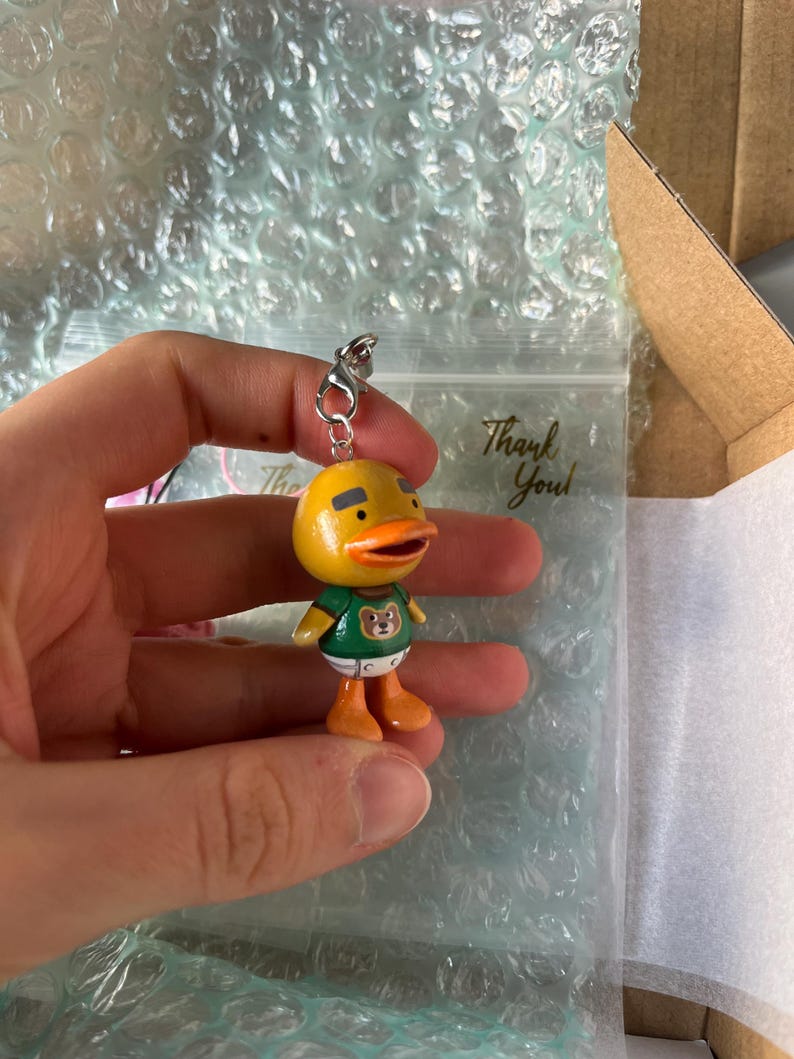 May include: A yellow duck character keychain with a green shirt featuring a brown bear graphic. The duck has a black beak and orange feet.