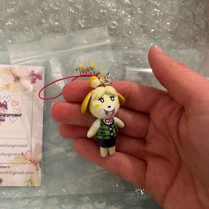 May include: A small, hand-painted figurine of a dog character wearing a green and white plaid vest and a black skirt. The figurine is attached to a red and white string.