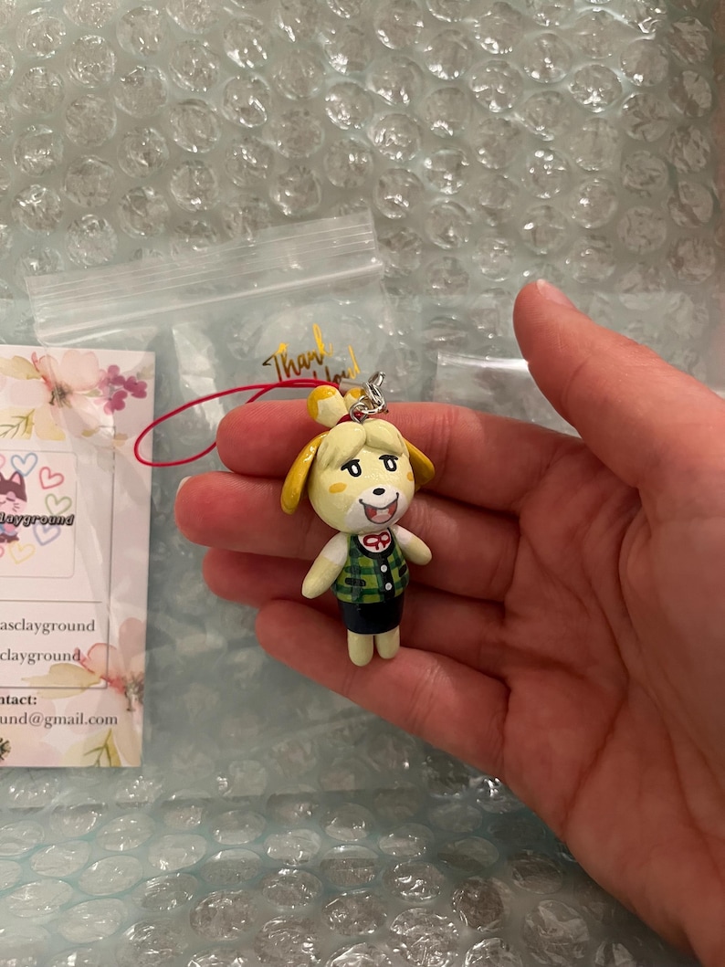 May include: A small, hand-painted figurine of a dog character wearing a green and white plaid vest and a black skirt. The figurine is attached to a red and white string.