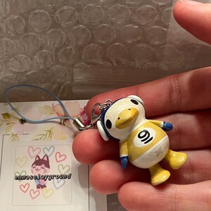 May include: A yellow duck character with blue accents and a white number 9 on its chest. The duck is wearing headphones and is a handmade charm. The charm is attached to a blue and white string.