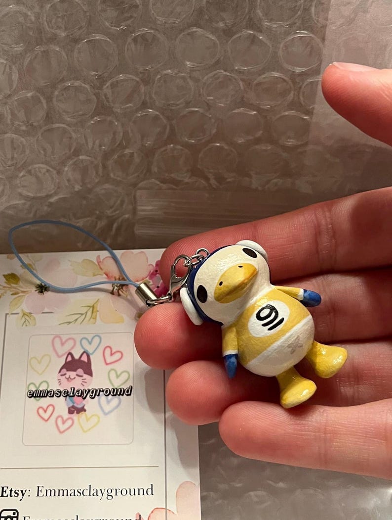 May include: A yellow duck character with blue accents and a white number 9 on its chest. The duck is wearing headphones and is a handmade charm. The charm is attached to a blue and white string.