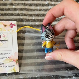 May include: A gray and white cat character with black eyes and glasses wearing a yellow sweater and blue jeans. The character is a charm with a silver loop for hanging.