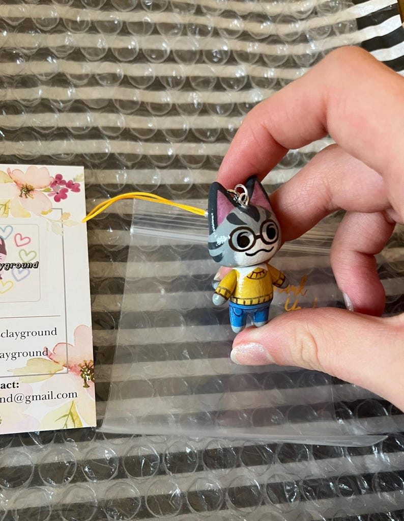 May include: A gray and white cat character with black eyes and glasses wearing a yellow sweater and blue jeans. The character is a charm with a silver loop for hanging.