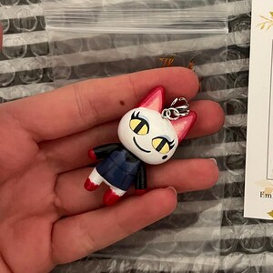 May include: A white and red cat figurine with yellow eyes and a black and blue shirt. The figurine is attached to a silver keychain.