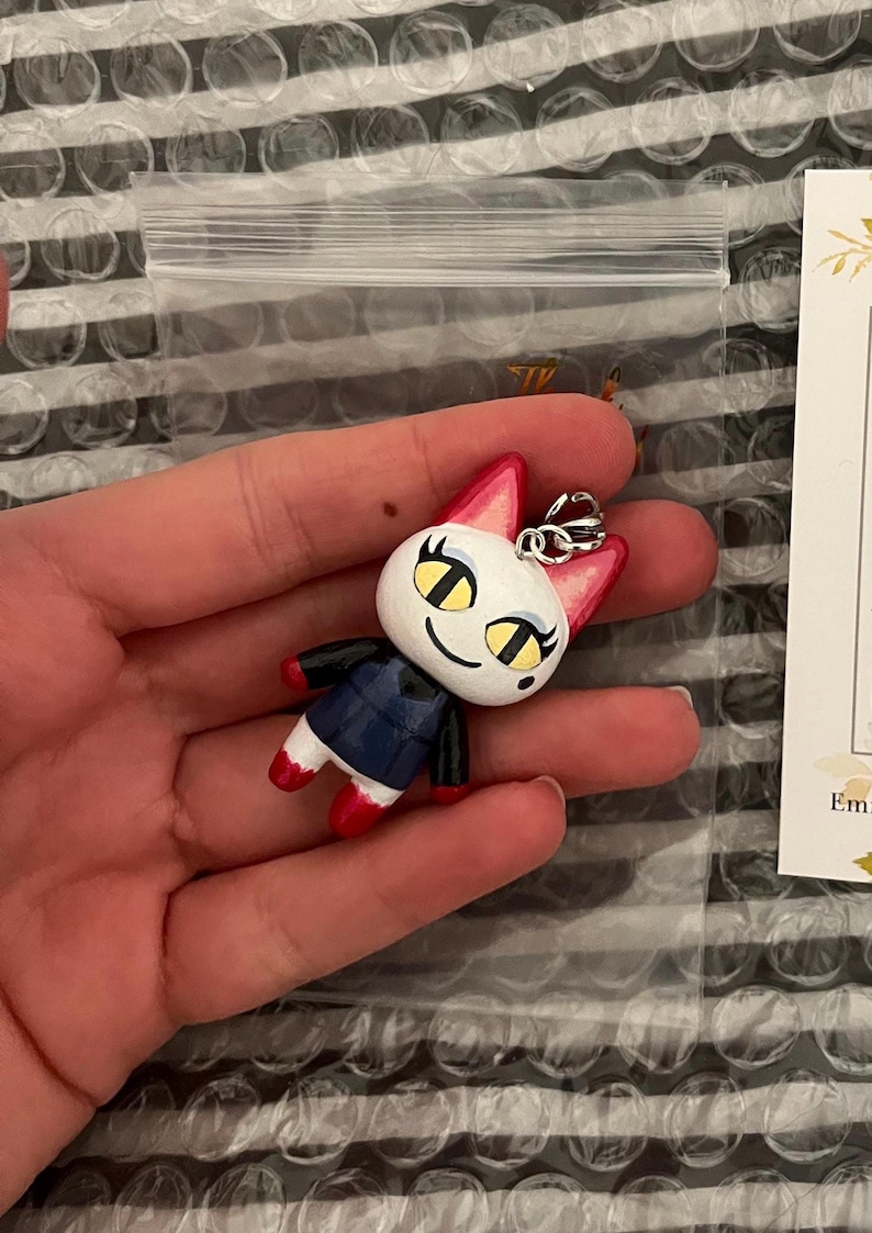May include: A white and red cat figurine with yellow eyes and a black and blue shirt. The figurine is attached to a silver keychain.