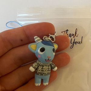 May include: A blue and white animal character keychain with a gray and white sweater. The keychain has a silver chain and a blue lanyard. The keychain is in a clear plastic bag with a sticker that says "Thank You".