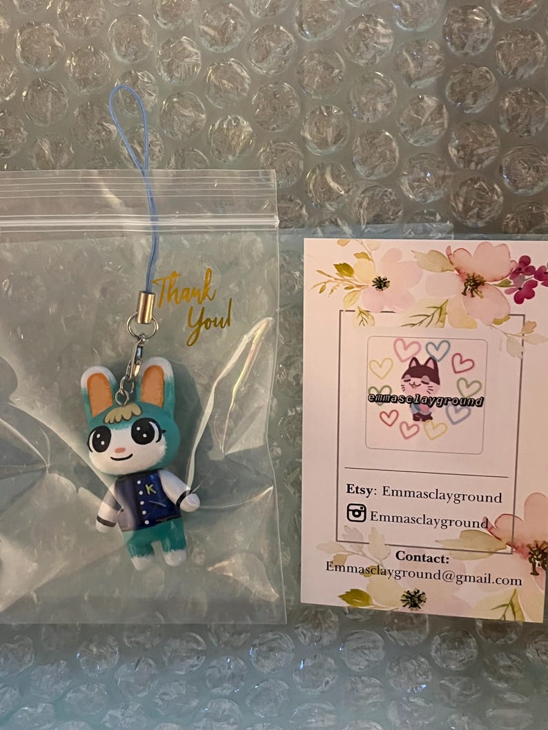 May include: A teal blue and white animal crossing character keychain with a blue jacket and a green hat. The character is holding a green sword. The keychain is in a clear plastic bag with a thank you note. The background is a white bubble wrap sheet.