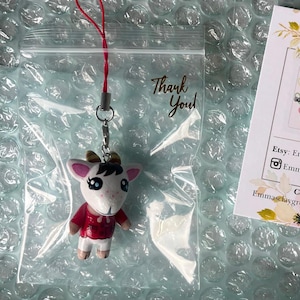 May include: A white goat character with brown horns and red clothing is attached to a red string and a silver chain. The character is a charm or keychain. The charm is in a clear plastic bag with a thank you note.