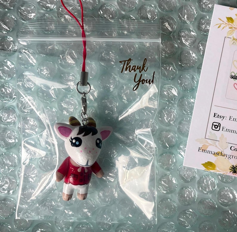 May include: A white goat character with brown horns and red clothing is attached to a red string and a silver chain. The character is a charm or keychain. The charm is in a clear plastic bag with a thank you note.