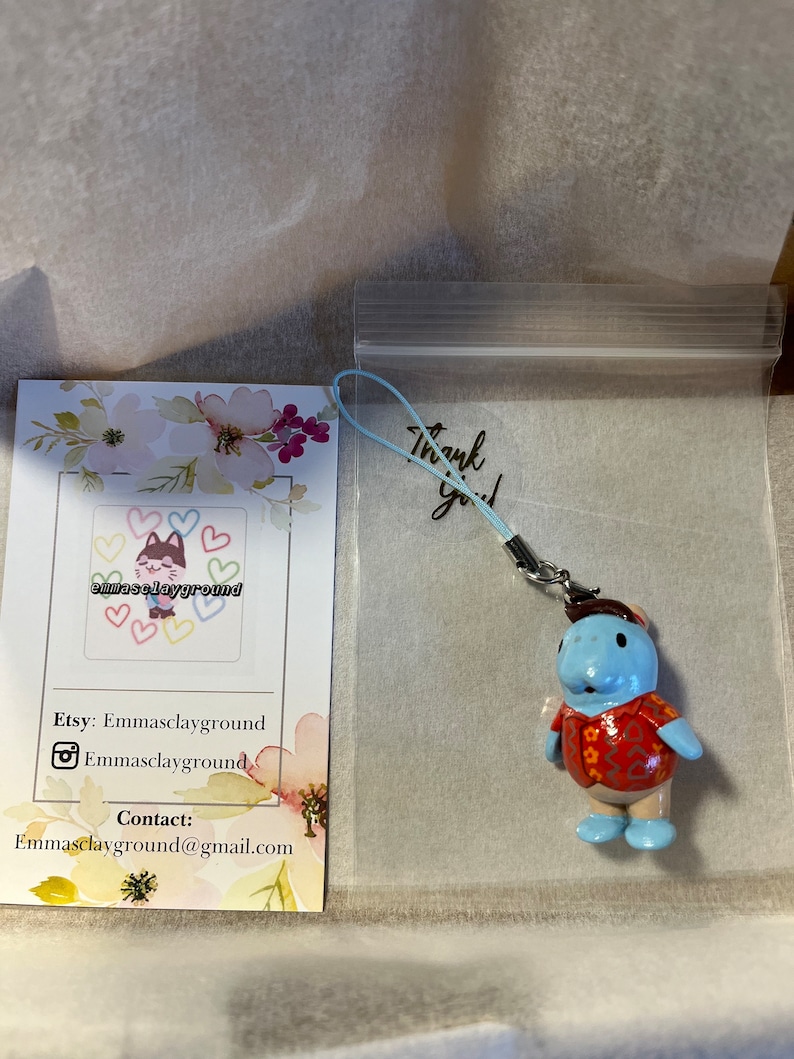 May include: A blue and red handmade polymer clay charm of a hippopotamus wearing a Hawaiian shirt. The charm is attached to a string and is packaged in a clear plastic bag with a thank you note.