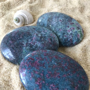 Puede incluir: Tres piedras lisas de forma ovalada con una base azul oscuro y un patrón moteado de rosa y morado. Las piedras están dispuestas en una pila sobre una superficie arenosa.
