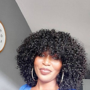 Può includere: Una donna con capelli scuri e ricci con frangia, indossa grandi orecchini a cerchio dorati e un top blu. Lo sfondo è un muro grigio chiaro e una porzione di un orologio.