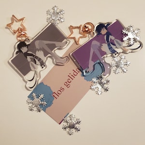 Puede incluir: Dos encantos de acrílico transparente con personajes de estilo anime con cabello largo y vestidos fluidos. Los encantos están unidos a llaveros de color dorado con encantos en forma de estrella. Los encantos están rodeados de encantos en forma de copo de nieve plateados y una tarjeta con el texto "flos gelida".