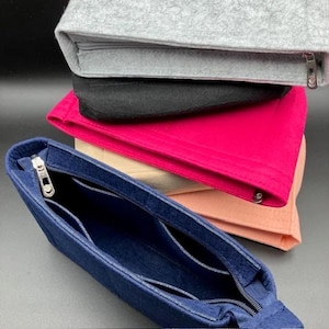 Puede incluir: Una pila de organizadores de bolsos de fieltro en varios colores: azul marino, rosa intenso, melocotón, negro y gris. Cada organizador tiene un cierre de cremallera y bolsillos interiores. Los organizadores están diseñados para caber dentro de un bolso.