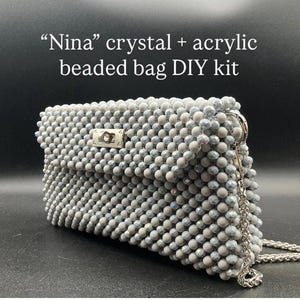 May include: A Nina crystal and acrylic beaded bag DIY kit featuring a rectangular handbag constructed from light gray, faceted beads. The bag includes a silver-toned metal turn-lock closure on the front flap and a matching silver-toned metal chain strap.
