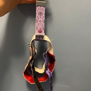 May include: A purple and black patterned key chain with a silver ring and a purple button. The key chain is made of fabric and has a black elastic band attached to it.