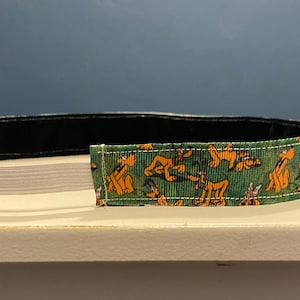 May include: A green and orange headband with a repeating pattern of cartoon dogs. The headband has a black fabric backing and a white elastic band.