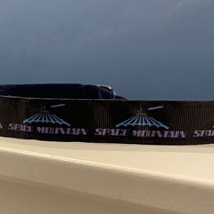 Könnte beinhalten: Schwarzes Band mit einem sich wiederholenden Muster des Space Mountain-Fahrgeschäftlogos und dem Text "Space Mountain".