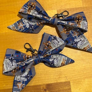 May include: Two bow ties with a Star Wars theme. The bow ties are made of fabric with a blue, gray, and white pattern featuring R2-D2 and C-3PO. Each bow tie has a silver-colored clasp.