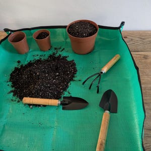 May include: A selection of gardening tools and supplies on a green mat. Includes a small shovel, a hand rake, and a trowel with wooden handles. There are also brown plastic pots filled with soil, and a pile of soil.